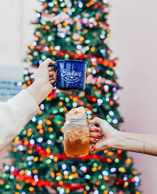 Hands hold a mug and a drinking jar in front of a Christmas tree with multicolored lights