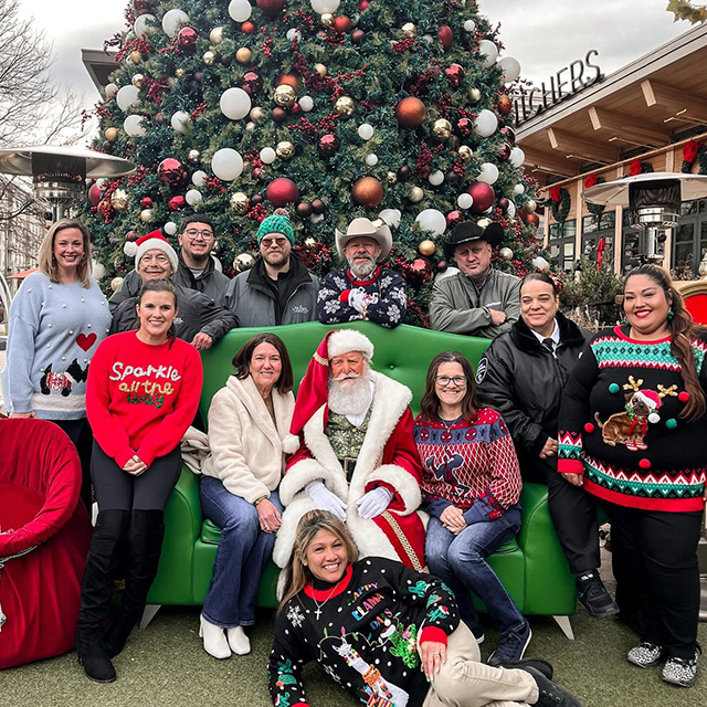 Santa sits on a couch in front of a large Christmas tree with a group of festively-dressed people