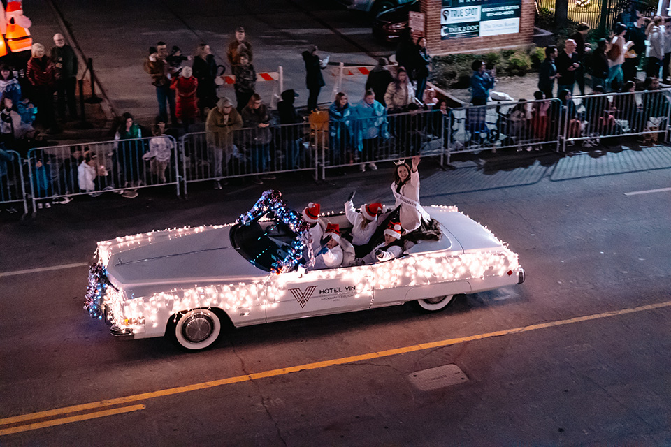 A classic car, decorated in Christmas lights, drives down the street while people in Santa hats wave to parade-goers