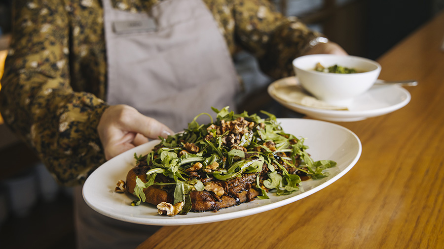 A beef dish and soup are held out by a person wearing an apron