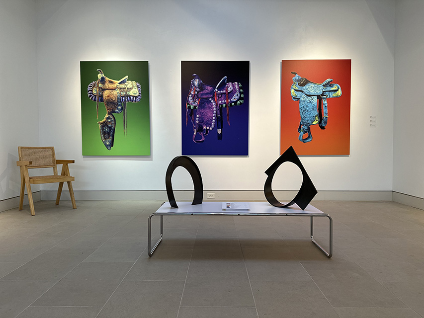 Art with saddles hangs on the wall of an art gallery, with sculptures on a table in front