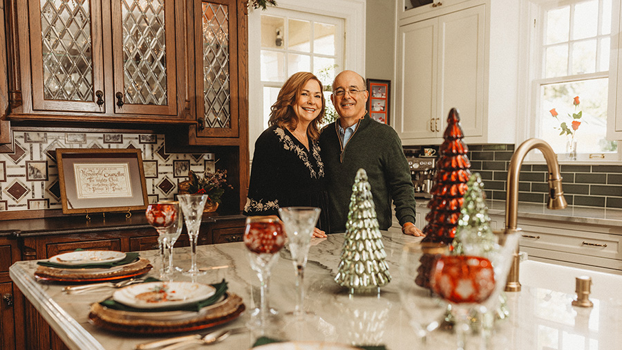 A man and woman stand in their beautiful kitchen, behind an island covered in festive Christmas decor and place settings