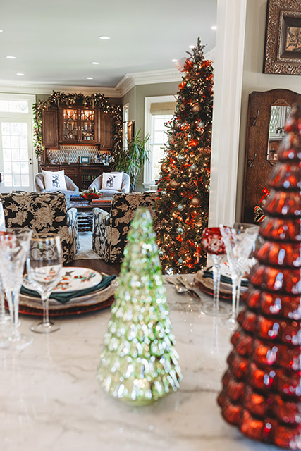 Glass christmas trees adorn a kitchen island, with a beautifully decorated living room and Christmas tree in the background