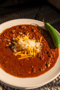 A bowl of red chili topped with cheese and a green pepper