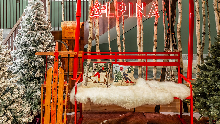 A neon "Alpina" sign above a wintery bench, sled and snowy trees.